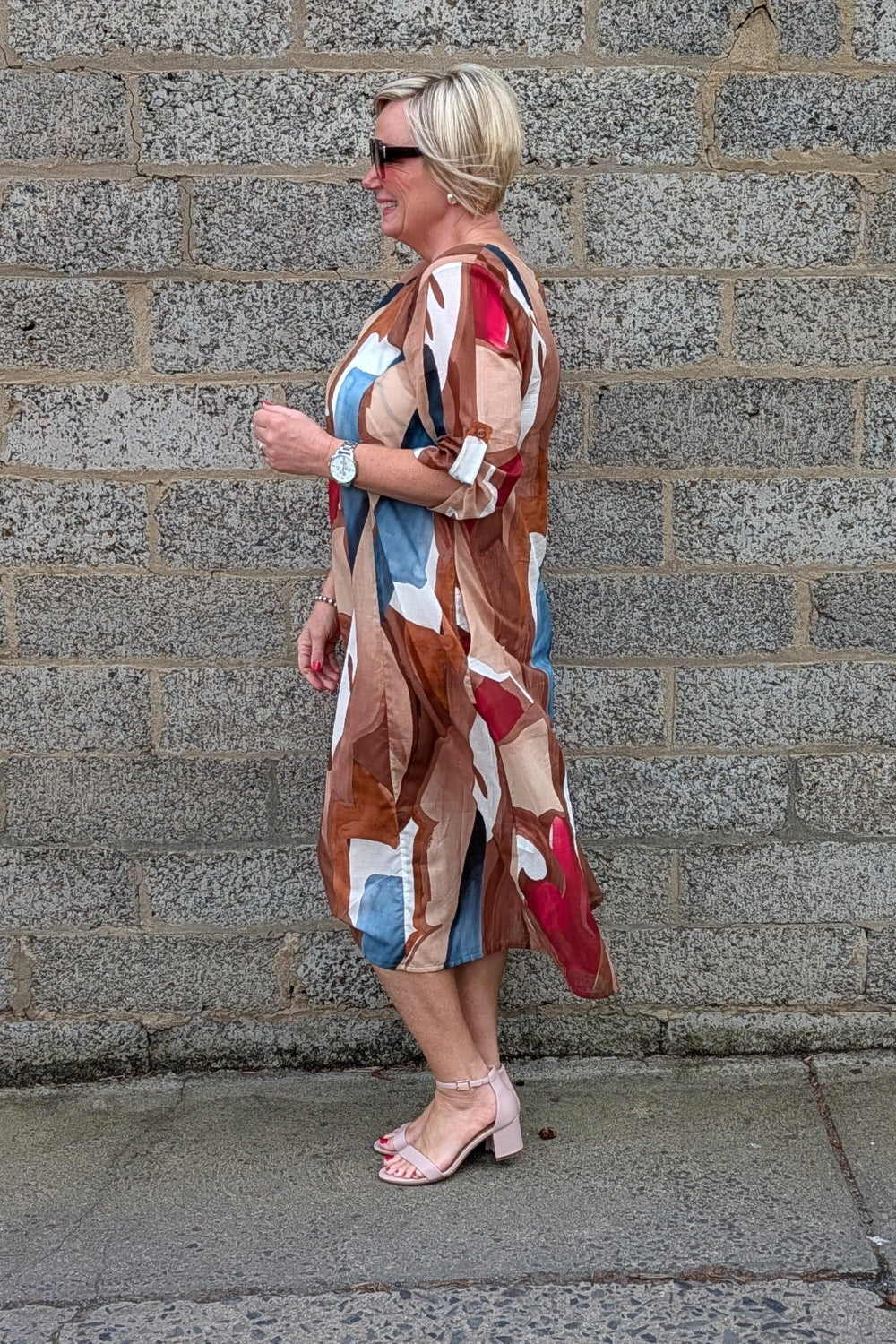 A woman wearing a long, abstract-patterned dress with brown, beige, blue, and red shapes, standing in profile against a gray stone wall.