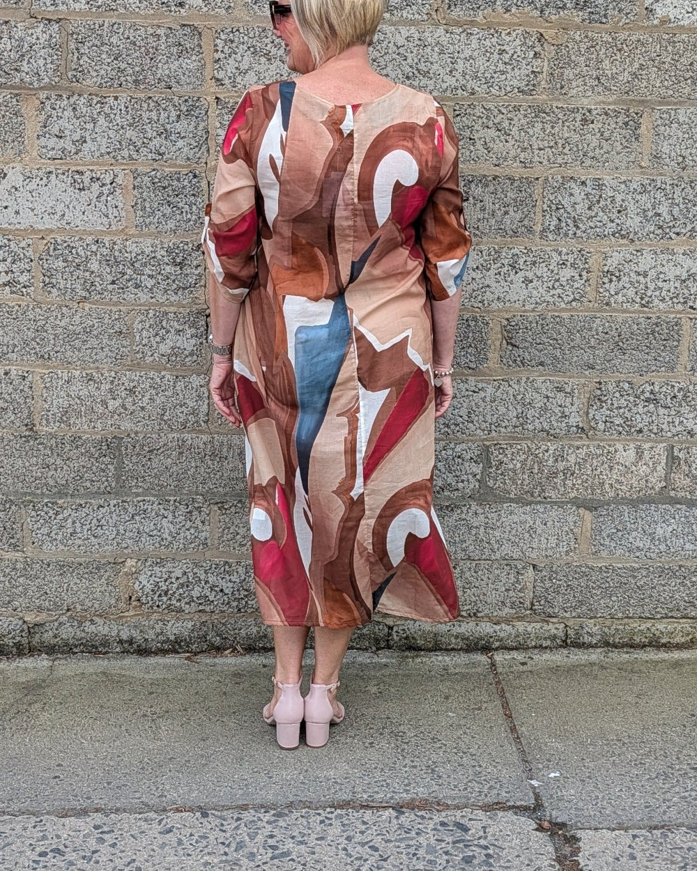 A woman with short blonde hair is shown from behind, wearing a long sleeved abstract print maxi dress with a mix of beige, brown, red, and blue tones. She is standing outdoors against a gray stone wall.