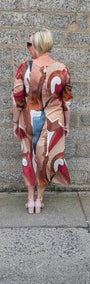 A woman with short blonde hair is shown from behind, wearing a long sleeved abstract print maxi dress with a mix of beige, brown, red, and blue tones. She is standing outdoors against a gray stone wall.