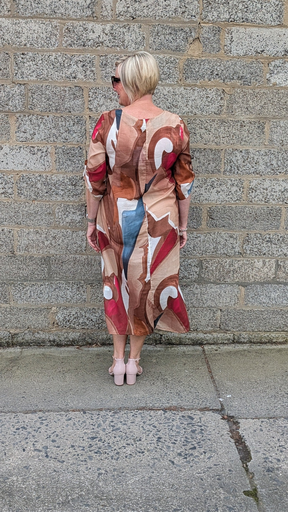 A woman with short blonde hair is shown from behind, wearing a long sleeved abstract print maxi dress with a mix of beige, brown, red, and blue tones. She is standing outdoors against a gray stone wall.