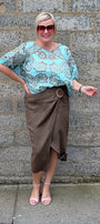 A woman posing in front of a gray stone wall, wearing a light blue patterned short sleeve blouse, a brown wrap midi skirt, and pink sandals.