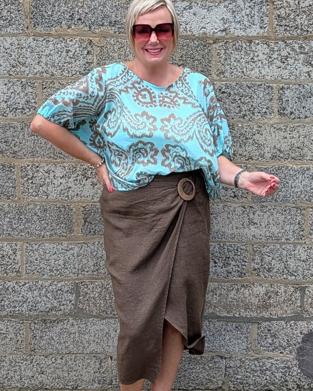 A woman modeling a light blue and brown paisley patterned blouse with a brown wrap skirt, standing against a gray stone wall.