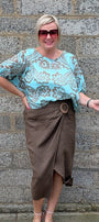 A woman modeling a light blue and brown paisley patterned blouse with a brown wrap skirt, standing against a gray stone wall.