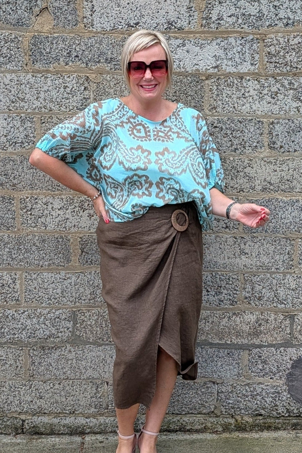 A woman modeling a light blue and brown paisley patterned blouse with a brown wrap skirt, standing against a gray stone wall.