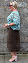 A woman wearing a turquoise and brown paisley patterned top with a brown midi skirt and light pink block heel sandals, standing in profile against a gray stone wall.