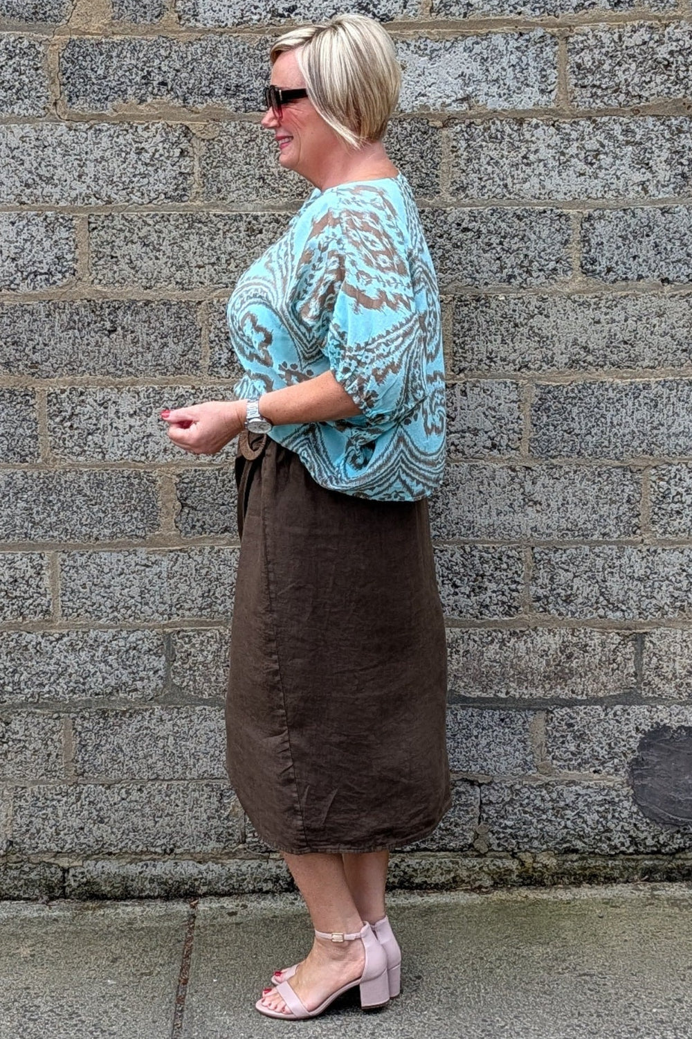 A woman wearing a turquoise and brown paisley patterned top with a brown midi skirt and light pink block heel sandals, standing in profile against a gray stone wall.