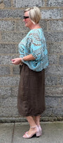 A woman stands in profile against a grey stone wall, wearing a light blue and white paisley patterned top, a brown midi skirt, and light pink heeled sandals.