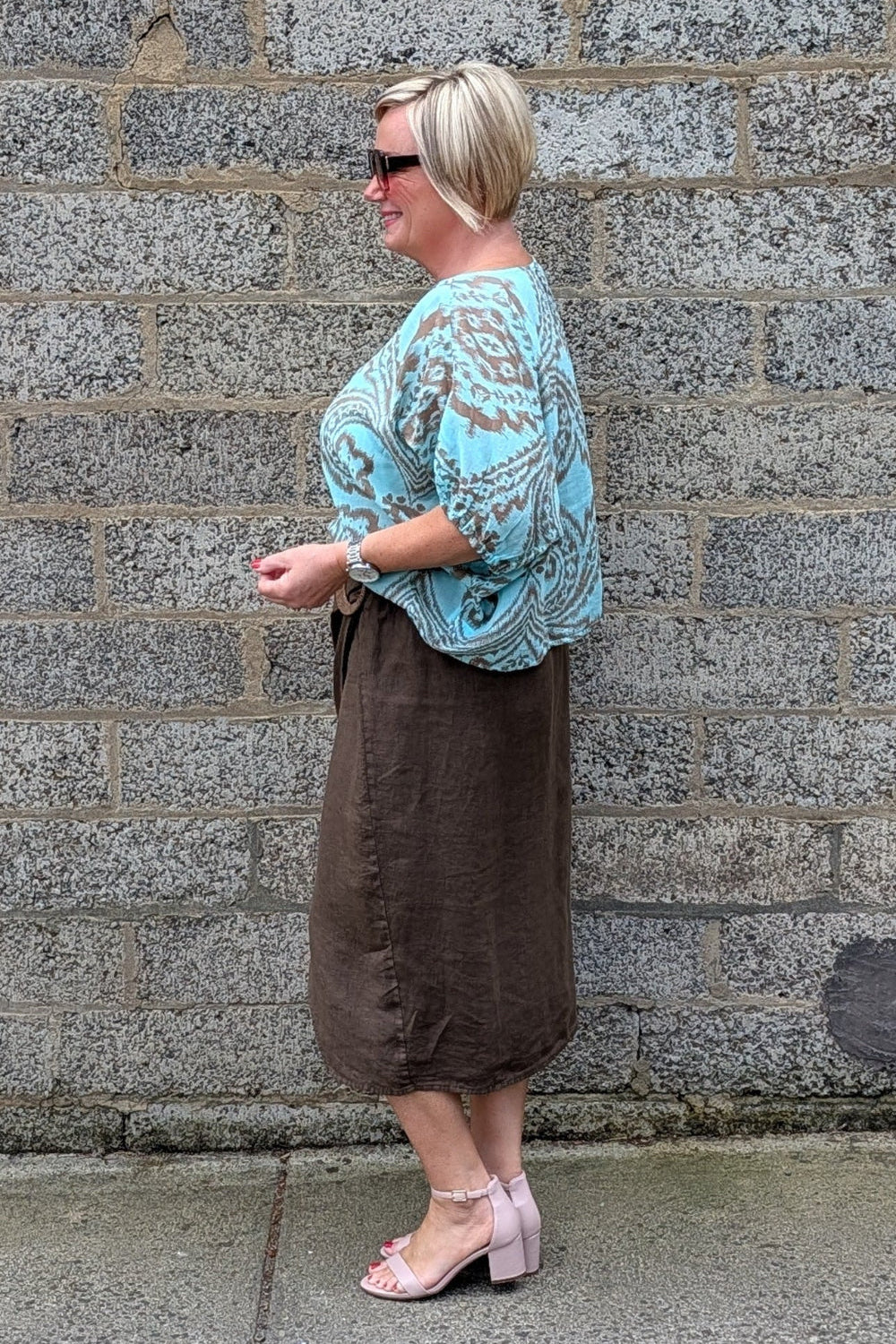 A woman stands in profile against a grey stone wall, wearing a light blue and white paisley patterned top, a brown midi skirt, and light pink heeled sandals.