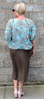 A woman viewed from behind wearing a light blue and brown paisley patterned blouse with puff sleeves and a brown midi skirt, standing against a gray stone wall.