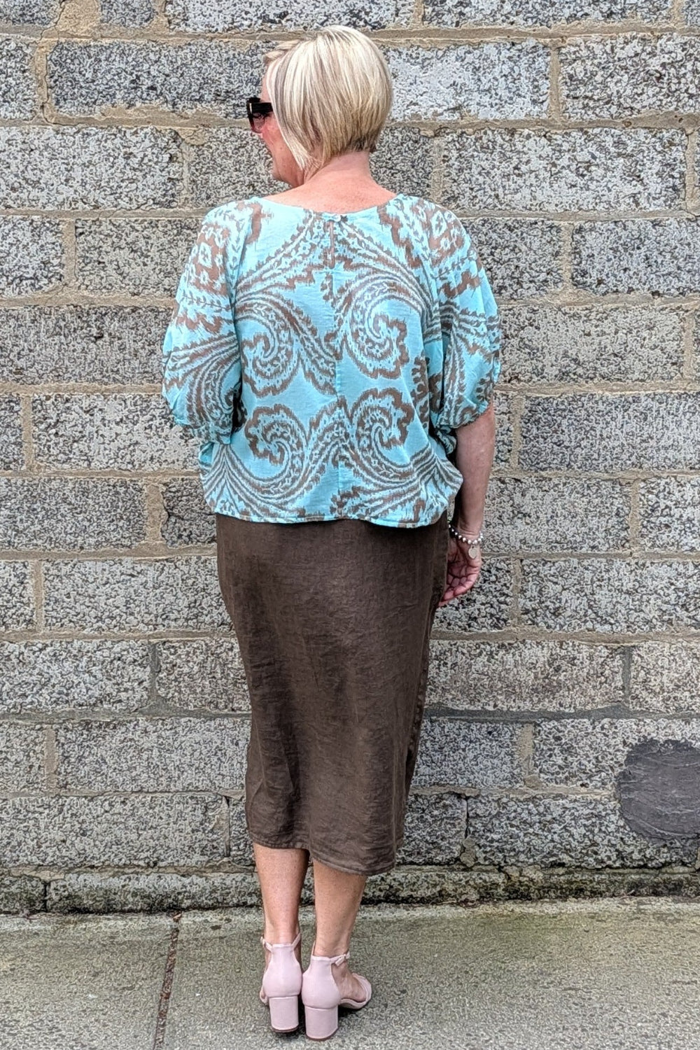 A woman viewed from behind wearing a light blue and brown paisley patterned blouse with puff sleeves and a brown midi skirt, standing against a gray stone wall.