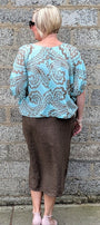 A woman viewed from behind wearing a light blue and brown paisley patterned blouse and a brown midi skirt, standing against a grey stone wall.