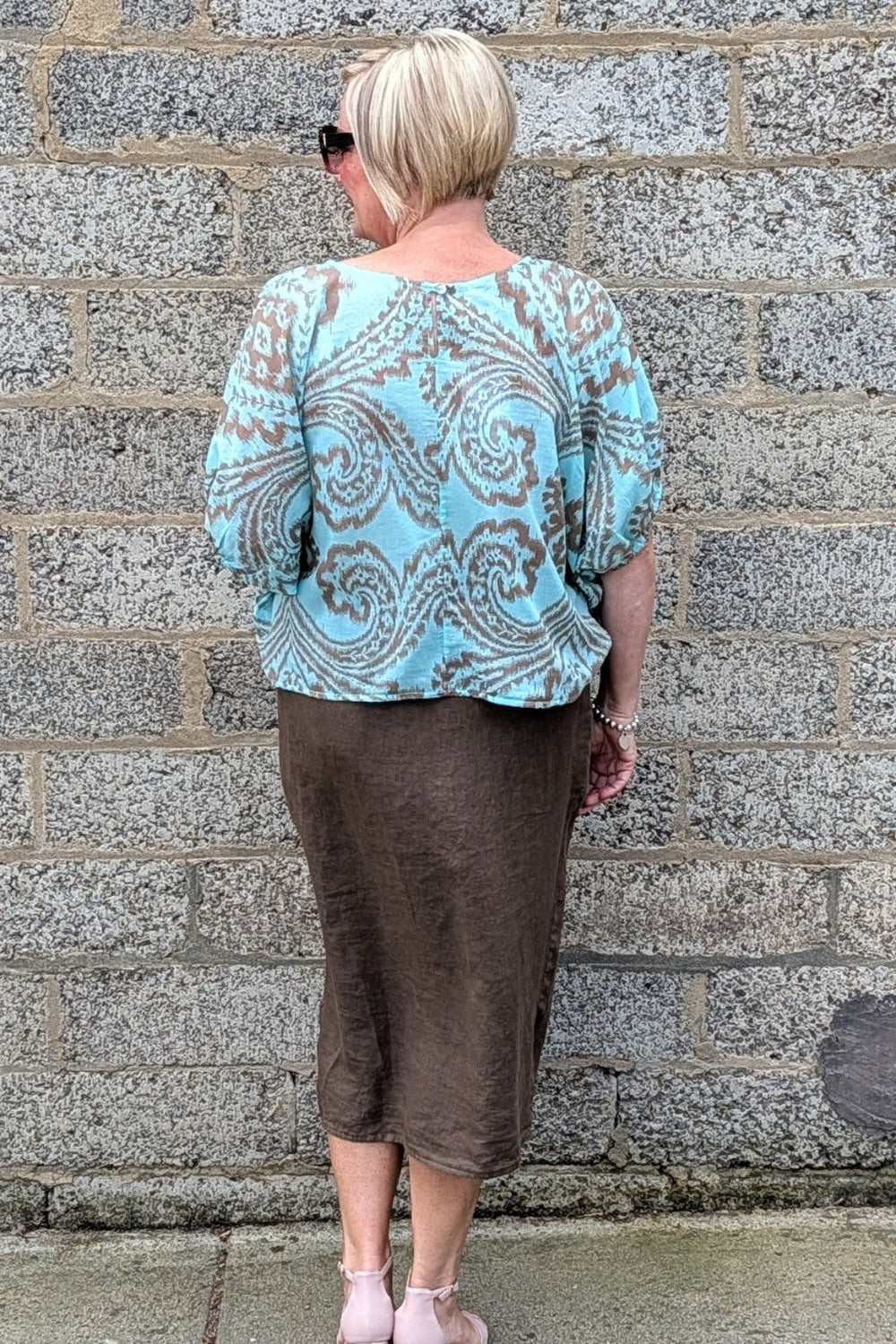 A woman viewed from behind wearing a light blue and brown paisley patterned blouse and a brown midi skirt, standing against a grey stone wall.