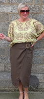 A woman wearing a light yellow and brown paisley patterned blouse and a brown wrap midi skirt with a large round brown belt, standing against a grey stone wall.