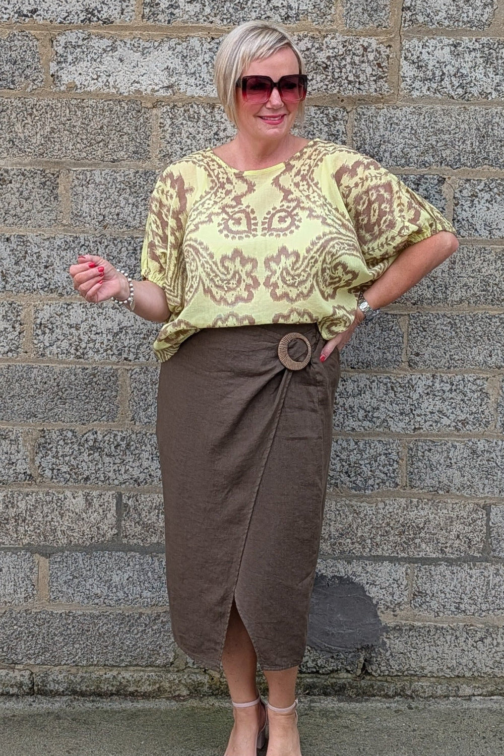 A woman wearing a light yellow and brown paisley patterned blouse and a brown wrap midi skirt with a large round brown belt, standing against a grey stone wall.