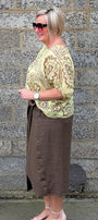 A woman stands in profile against a grey stone wall, wearing a light yellow green and brown paisley patterned top, a brown midi skirt, and light pink heeled sandals.