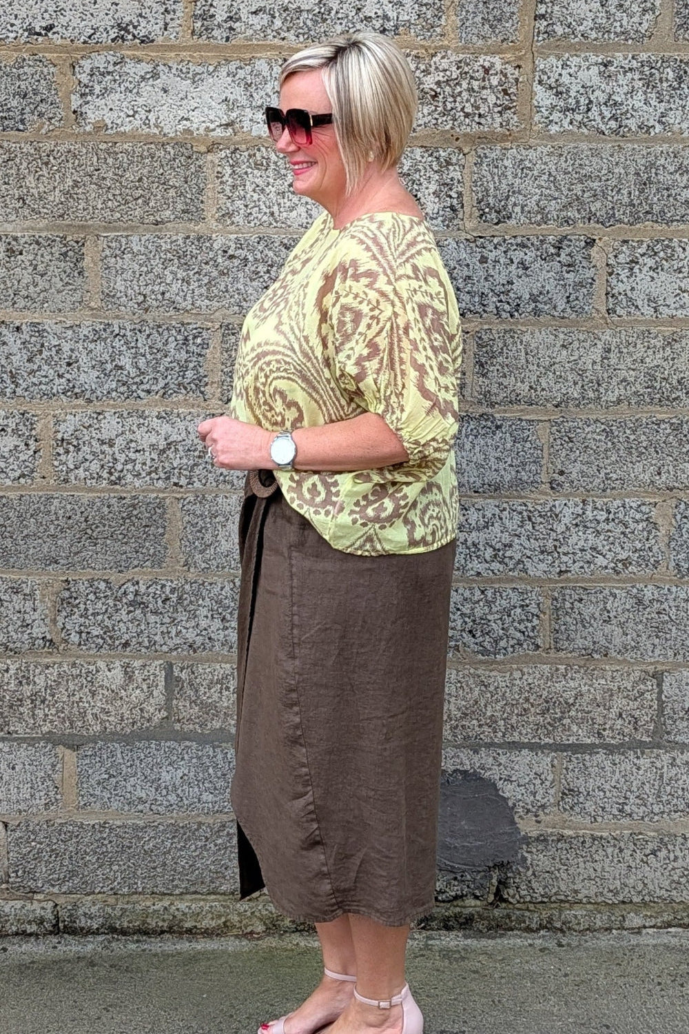 A woman stands in profile against a grey stone wall, wearing a light yellow green and brown paisley patterned top, a brown midi skirt, and light pink heeled sandals.