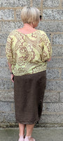 A woman viewed from behind stands against a gray stone wall, wearing a light yellow green and brown paisley patterned top with a brown midi skirt and light pink heels.