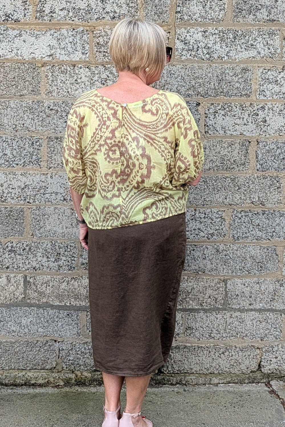 A woman viewed from behind stands against a gray stone wall, wearing a light yellow green and brown paisley patterned top with a brown midi skirt and light pink heels.