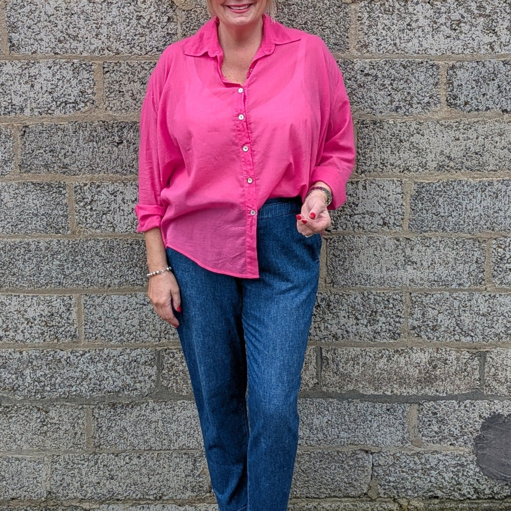 A woman wearing a bright pink button up shirt, blue jeans, and pink sandals stands in front of a gray stone wall.