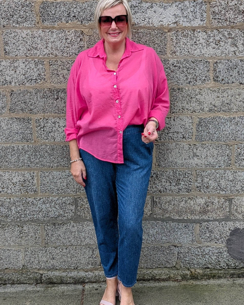 A woman wearing a bright pink button up shirt, blue jeans, and pink sandals stands in front of a gray stone wall.