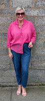 A woman wearing a bright pink button up shirt, blue jeans, and pink sandals stands in front of a gray stone wall.