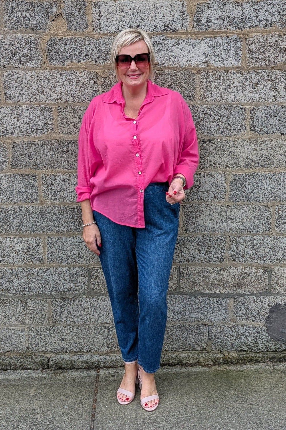 A woman wearing a bright pink button up shirt, blue jeans, and pink sandals stands in front of a gray stone wall.