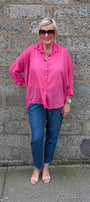 A woman wearing a bright pink button down shirt, blue jeans, and pink sandals stands in front of a gray stone wall.