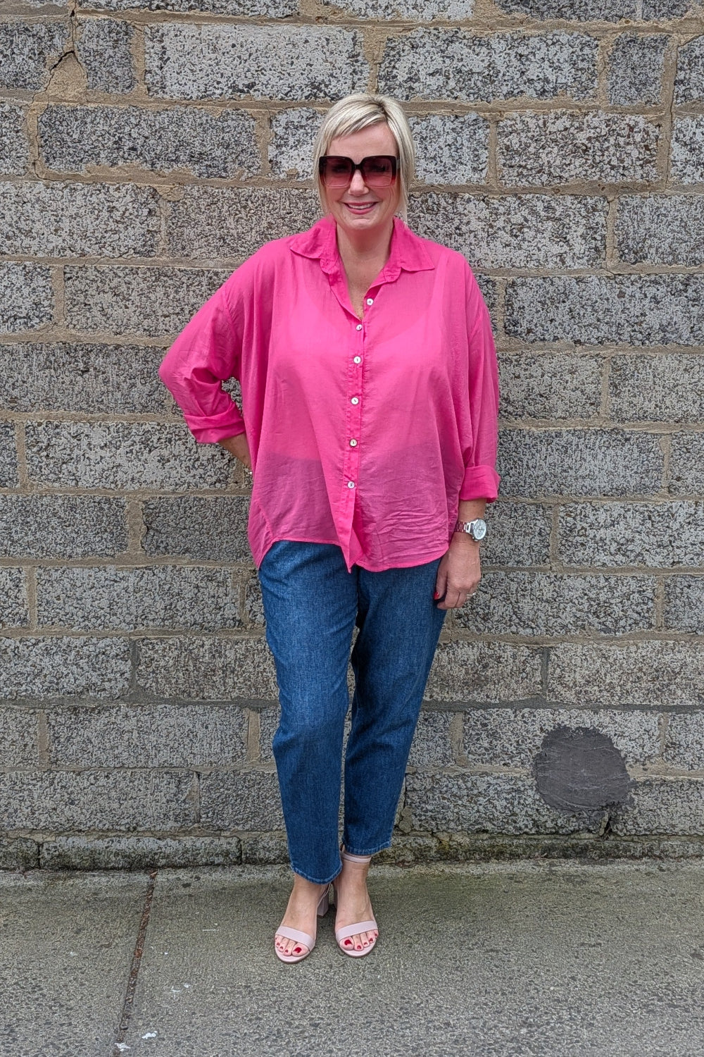 A woman wearing a bright pink button down shirt, blue jeans, and pink sandals stands in front of a gray stone wall.