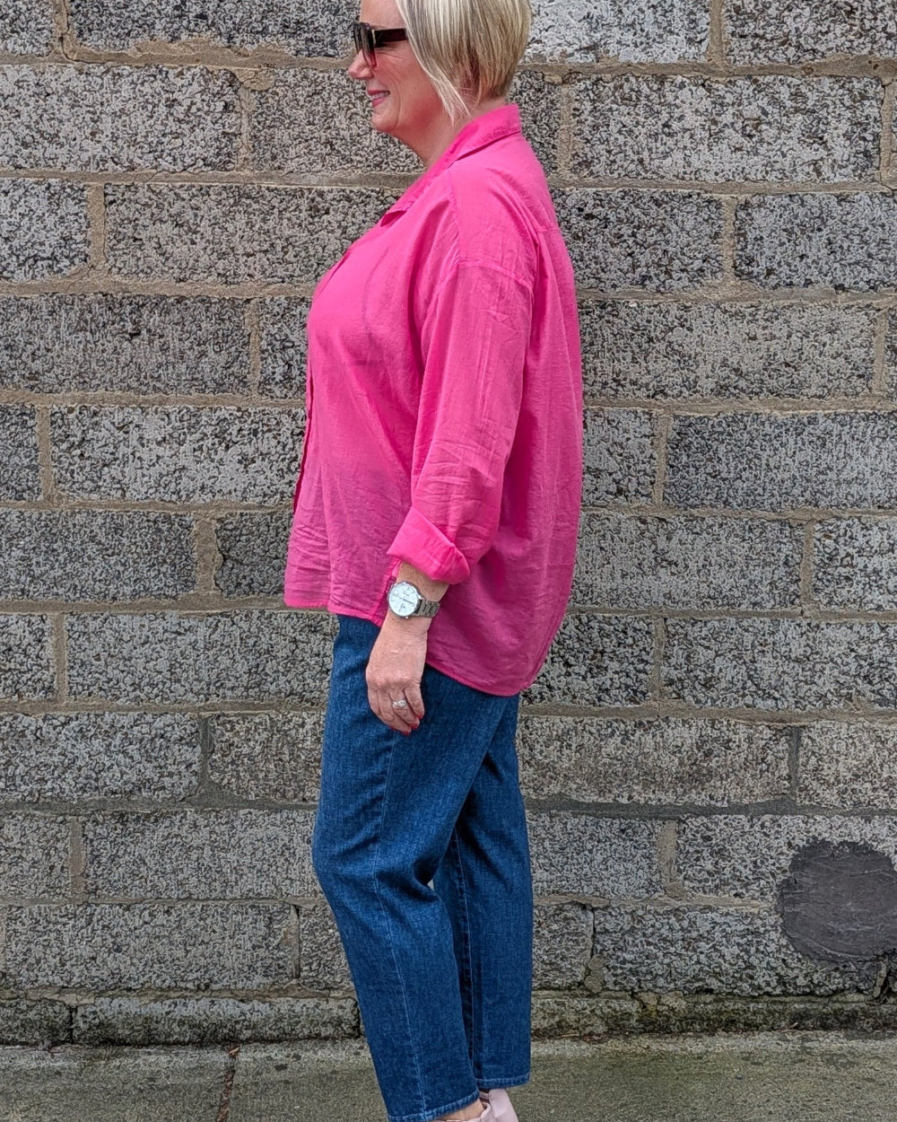 A woman in a bright pink button up shirt, blue jeans, and pink block heel sandals stands in profile against a gray stone wall.