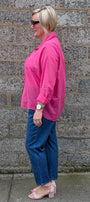 A woman in a bright pink button up shirt, blue jeans, and pink block heel sandals stands in profile against a gray stone wall.