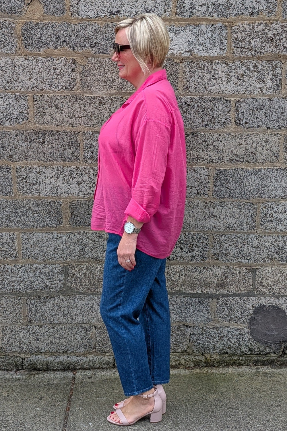 A woman in a bright pink button up shirt, blue jeans, and pink block heel sandals stands in profile against a gray stone wall.