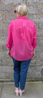 A woman viewed from behind wearing a bright pink button up shirt, blue jeans, and light pink heels, standing against a gray stone wall.