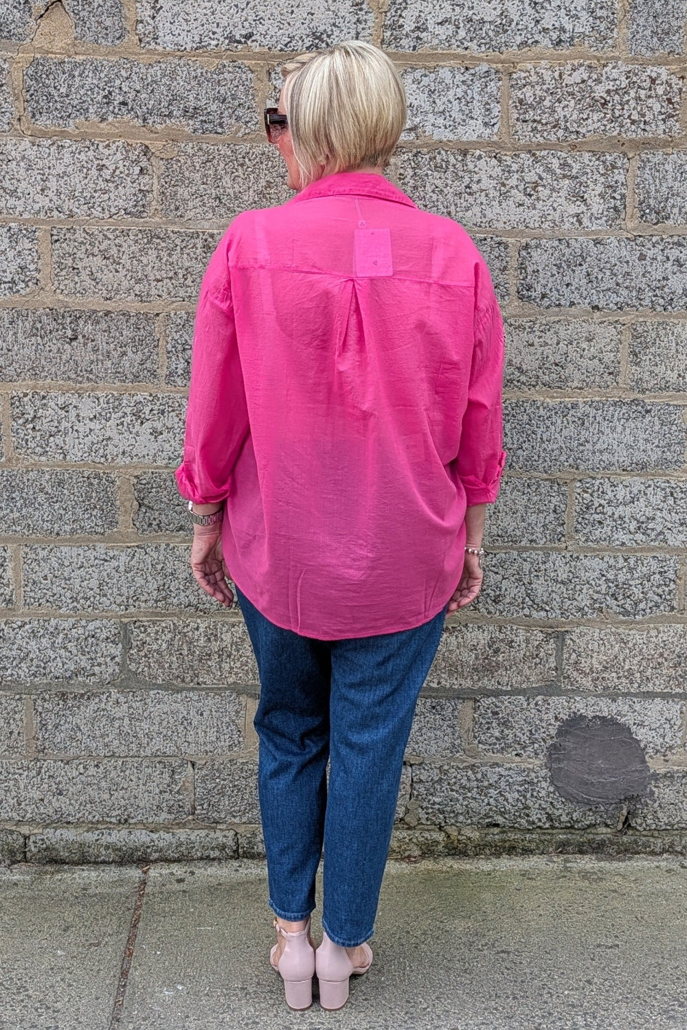 A woman viewed from behind wearing a bright pink button up shirt, blue jeans, and light pink heels, standing against a gray stone wall.