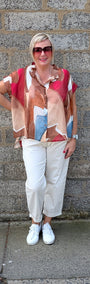 A woman stands against a gray stone wall wearing a short sleeve button up shirt with an abstract geometric print in red, beige, brown, and light blue, paired with white cropped pants and white sneakers.