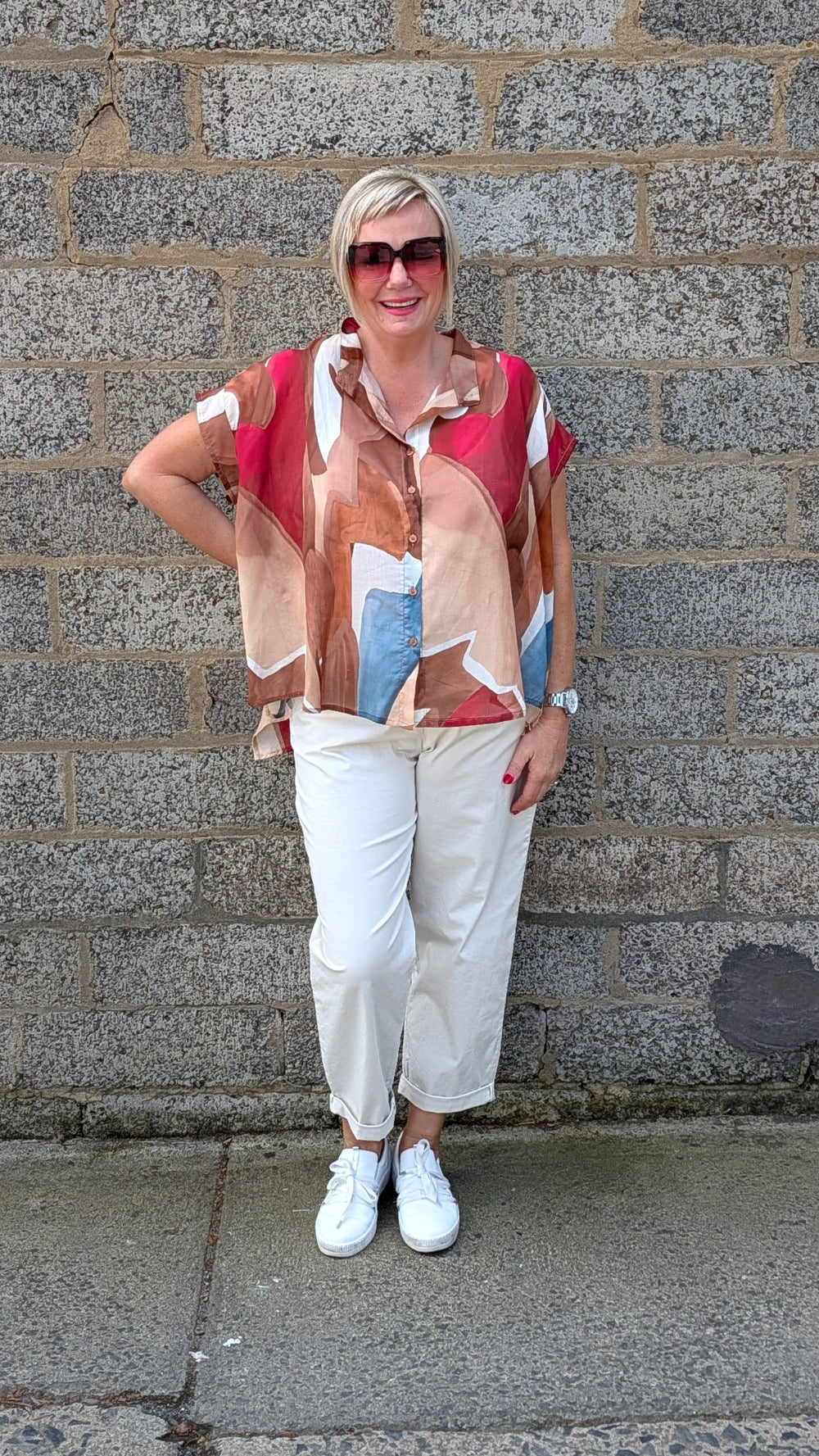 A woman stands against a gray stone wall wearing a short sleeve button up shirt with an abstract geometric print in red, beige, brown, and light blue, paired with white cropped pants and white sneakers.