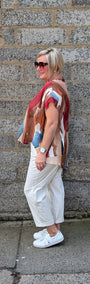 A woman stands in profile against a grey stone wall, wearing a multicolour patchwork knit top with white cropped pants and white sneakers.
