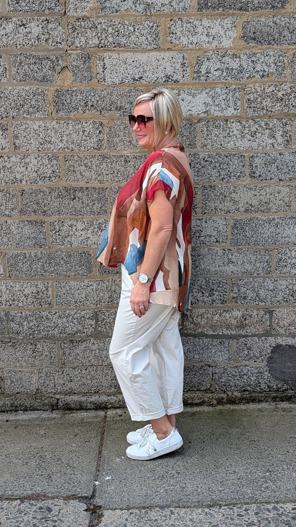 A woman stands in profile against a grey stone wall, wearing a multicolour patchwork knit top with white cropped pants and white sneakers.
