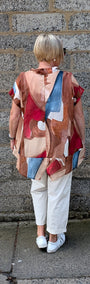 A woman viewed from behind stands against a grey stone wall wearing a loose abstract print top with red, beige, brown, and blue shapes, white pants, and white sneakers.
