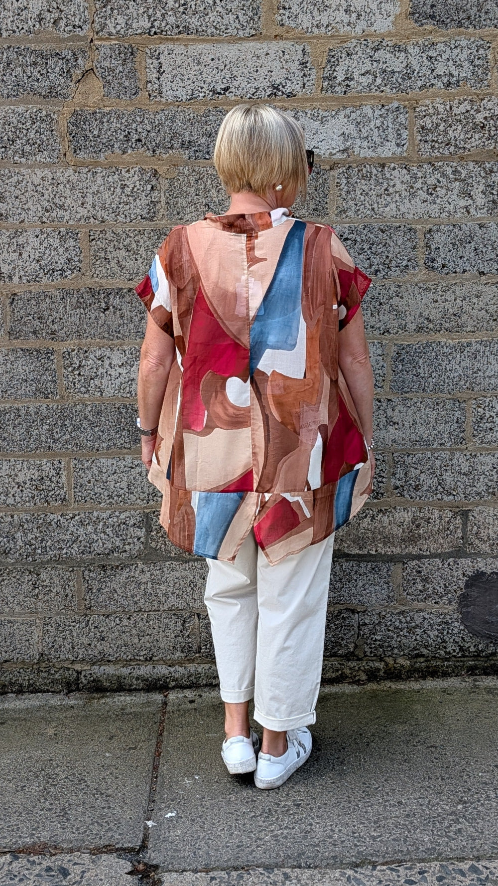 A woman viewed from behind stands against a grey stone wall wearing a loose abstract print top with red, beige, brown, and blue shapes, white pants, and white sneakers.