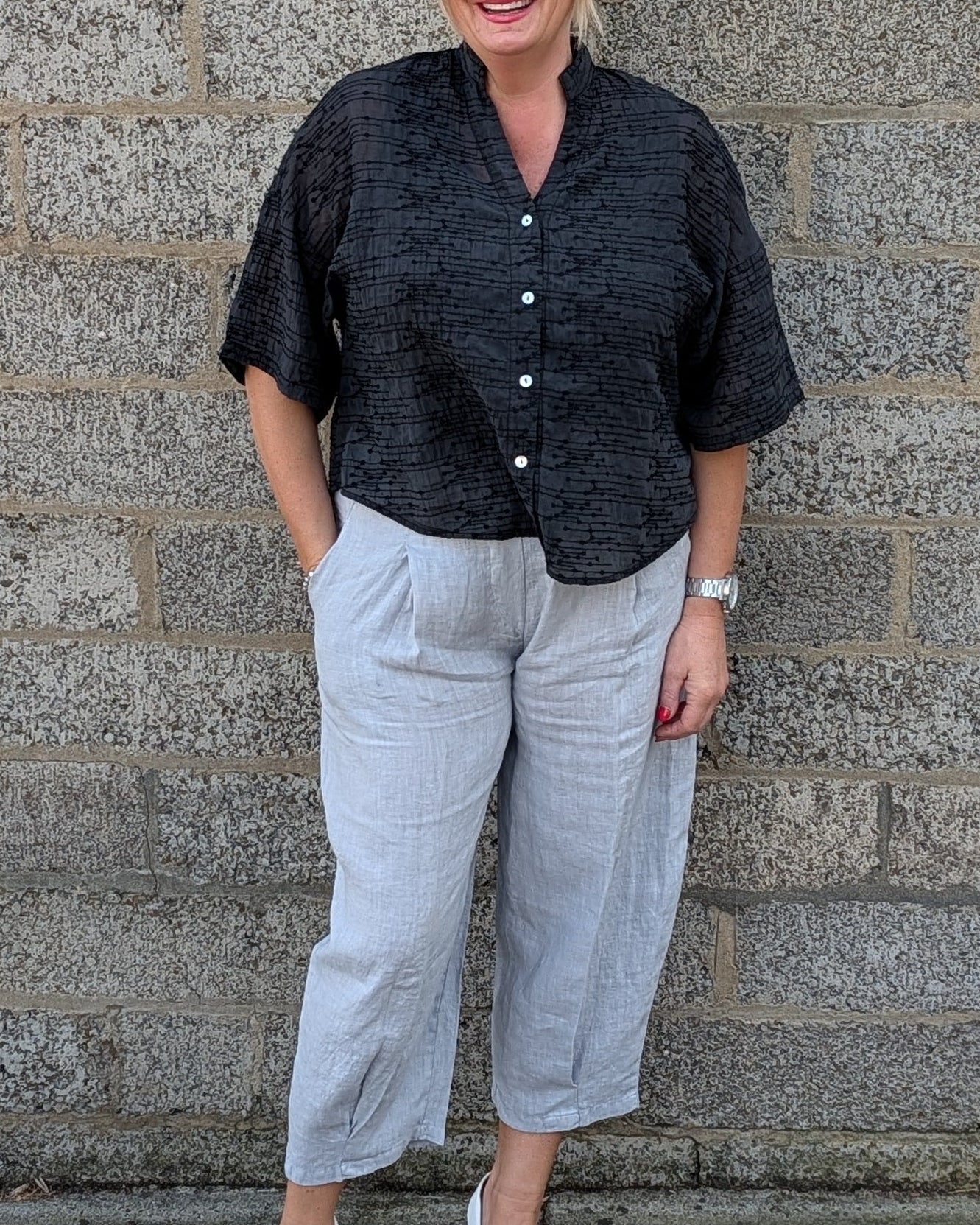 A woman stands against a gray stone wall wearing a black short sleeve button up shirt, light gray cropped trousers, and white pointed toe heels.