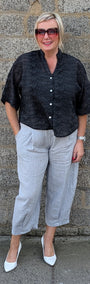 A woman stands against a gray stone wall wearing a black short sleeve button up shirt, light gray cropped trousers, and white pointed toe heels.