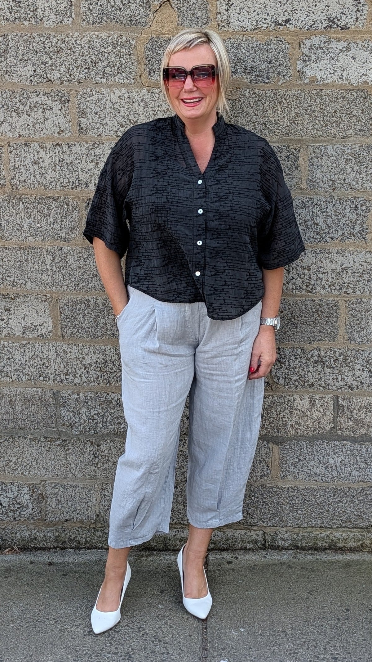 A woman stands against a gray stone wall wearing a black short sleeve button up shirt, light gray cropped trousers, and white pointed toe heels.