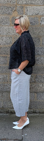 A woman stands in profile against a gray stone wall, wearing a black textured short sleeve top, a light gray midi skirt, and white pointed flats.