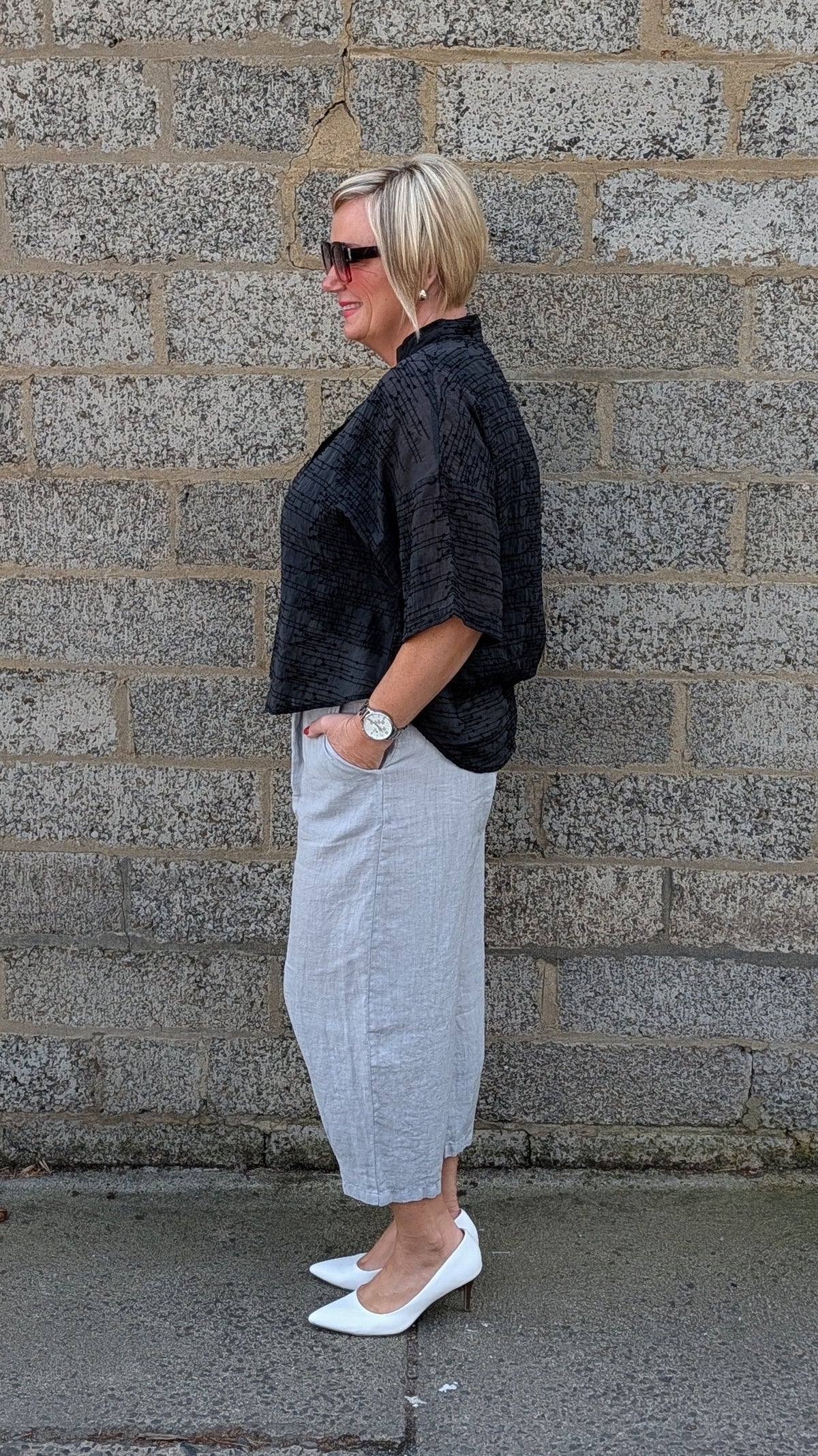 A woman stands in profile against a gray stone wall, wearing a black textured short sleeve top, a light gray midi skirt, and white pointed flats.