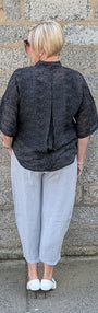 A person viewed from behind stands against a gray stone wall, wearing a dark gray textured short-sleeve top, light gray cropped pants, and white shoes.