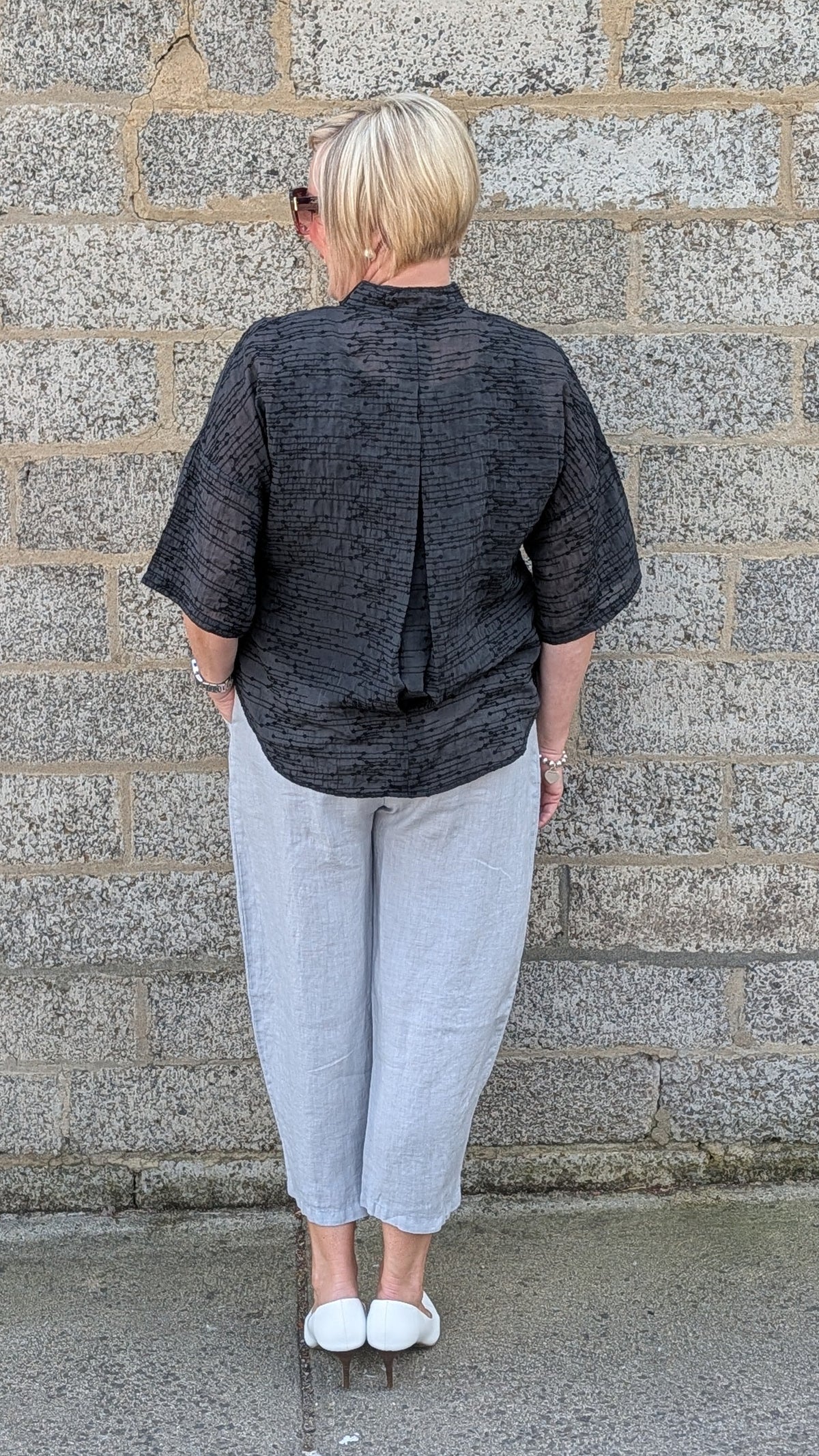 A person viewed from behind stands against a gray stone wall, wearing a dark gray textured short-sleeve top, light gray cropped pants, and white shoes.
