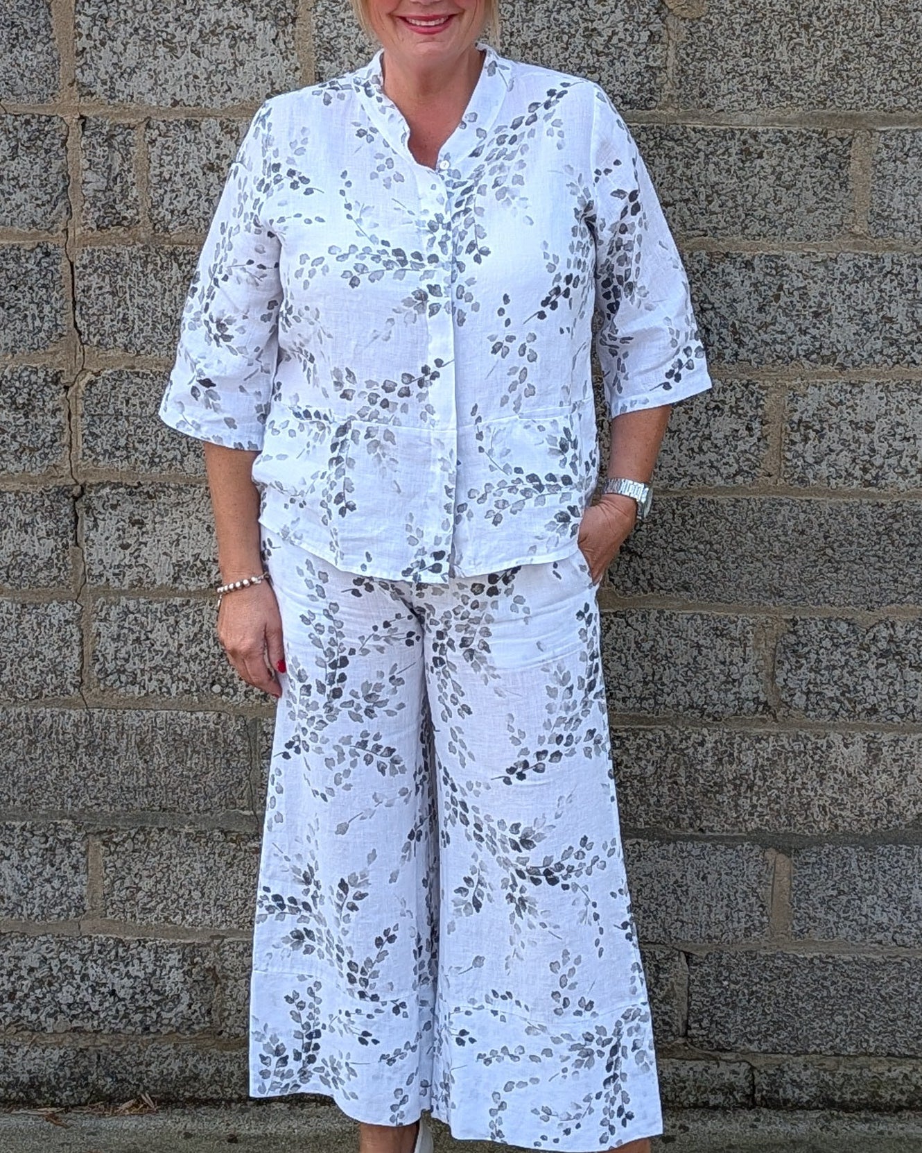 Front view white two piece matching set with a black floral pattern, consisting of a short sleeve button front top and wide leg pants, paired with white pointed shoes.
