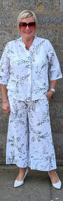 Front view white two piece matching set with a black floral pattern, consisting of a short sleeve button front top and wide leg pants, paired with white pointed shoes.