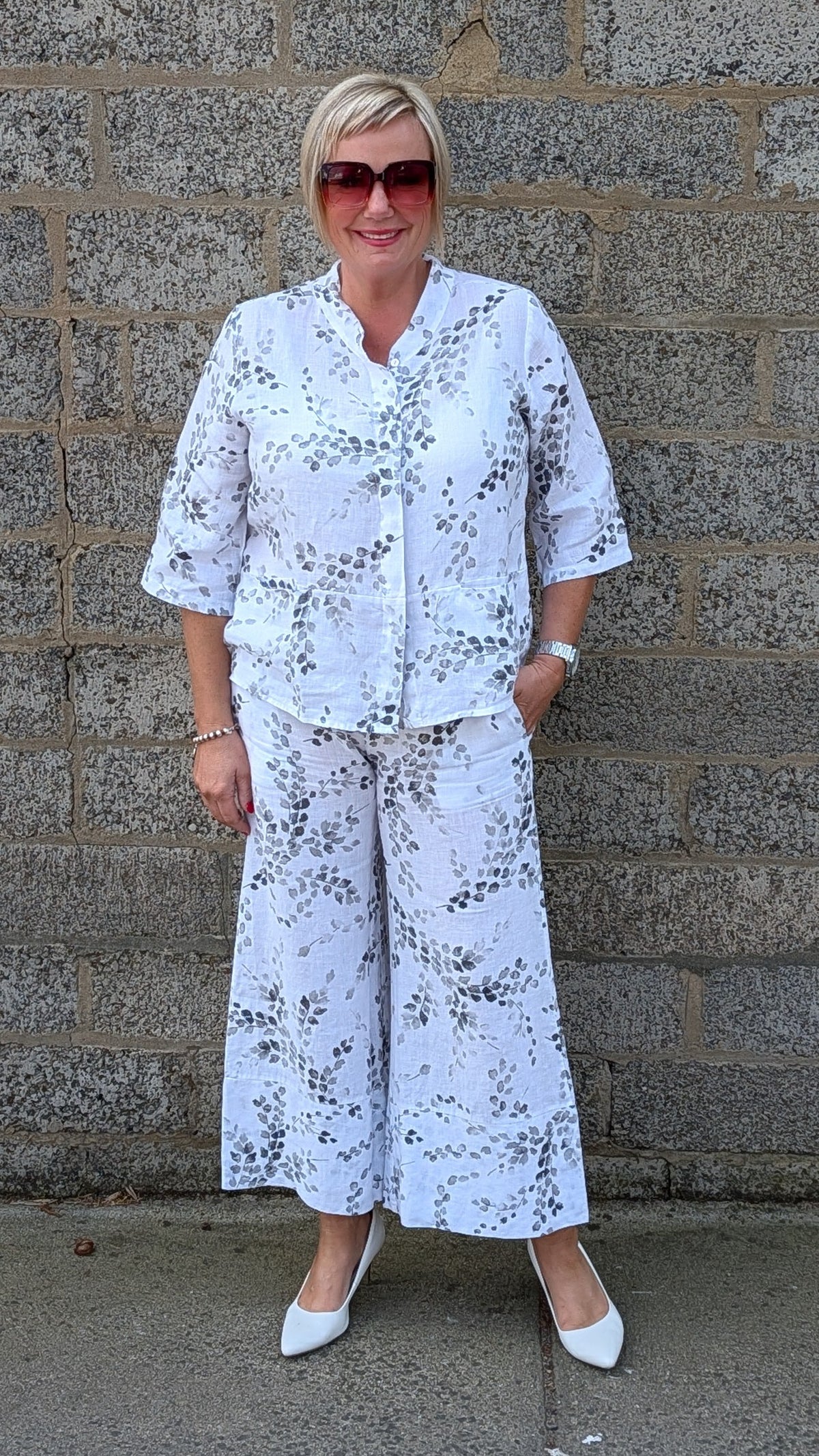 Front view white two piece matching set with a black floral pattern, consisting of a short sleeve button front top and wide leg pants, paired with white pointed shoes.
