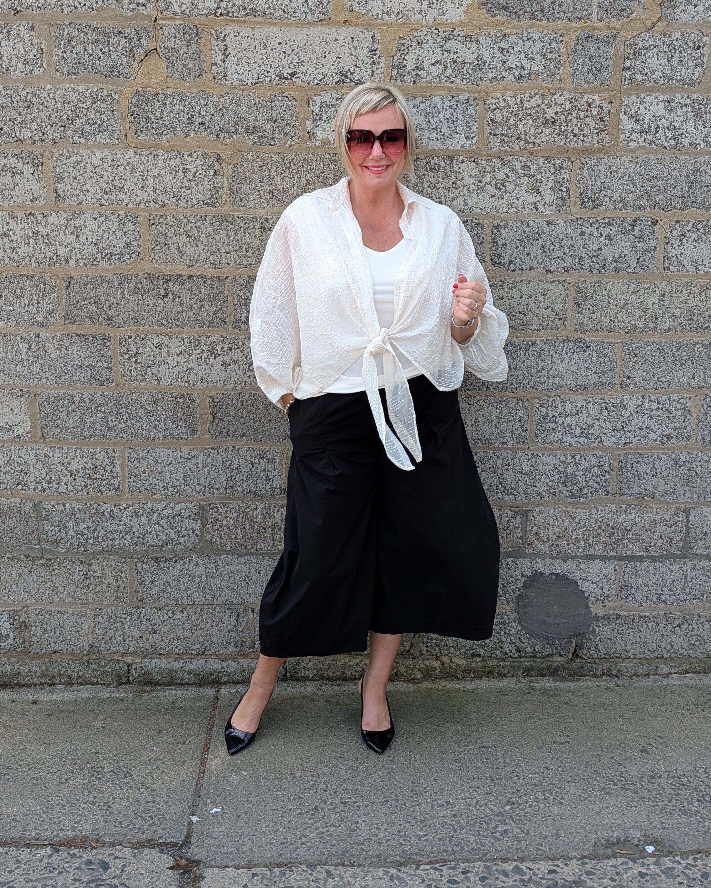 A woman stands against a gray stone wall wearing a white long sleeve tie front blouse, black wide leg cropped pants, and black pointed flats.